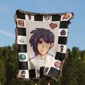 May include: A woven throw blanket with a pixelated design featuring a character from a video game. The character is a young person with blue hair and a white shirt. The blanket has a black and white checkered background with various pixelated icons.