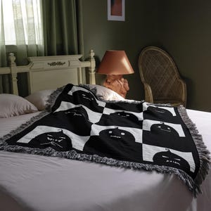 May include: A white bed with a black and white throw blanket featuring a repeating pattern of black jack-o'-lantern faces. The blanket has a fringe border.
