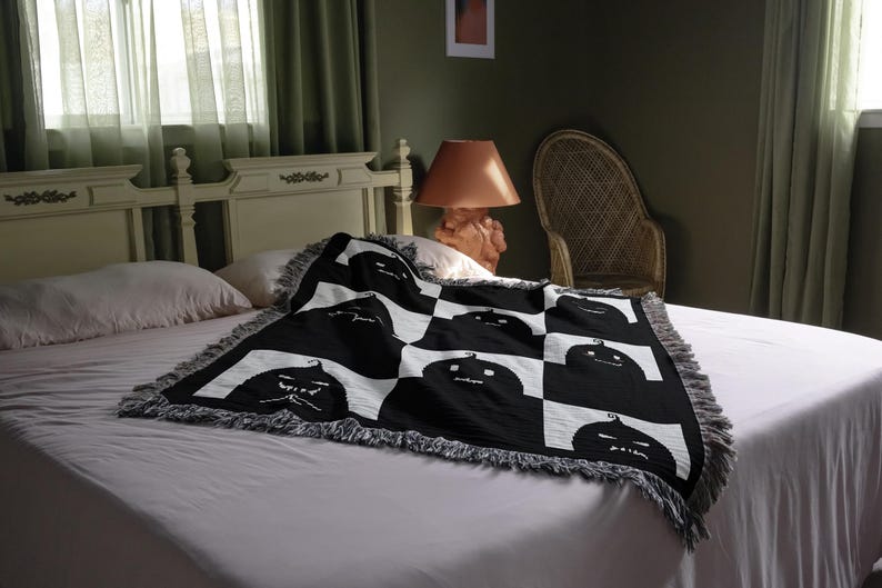 May include: A white bed with a black and white throw blanket featuring a repeating pattern of black jack-o'-lantern faces. The blanket has a fringe border.