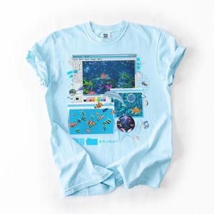 May include: Light blue t-shirt featuring a retro computer graphic with an aquarium scene. The graphic includes a dolphin, various fish, and the Japanese text "テクノロジー". The shirt has a classic crew neck and short sleeves.