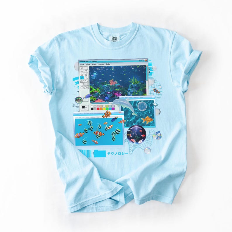 May include: Light blue t-shirt featuring a retro computer graphic with an aquarium scene. The graphic includes a dolphin, various fish, and the Japanese text "テクノロジー". The shirt has a classic crew neck and short sleeves.