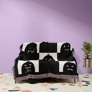 May include: Black and white woven throw blanket with a checkered pattern and a repeating design of black pumpkin faces. The blanket is draped over a black sofa with a fringe border.