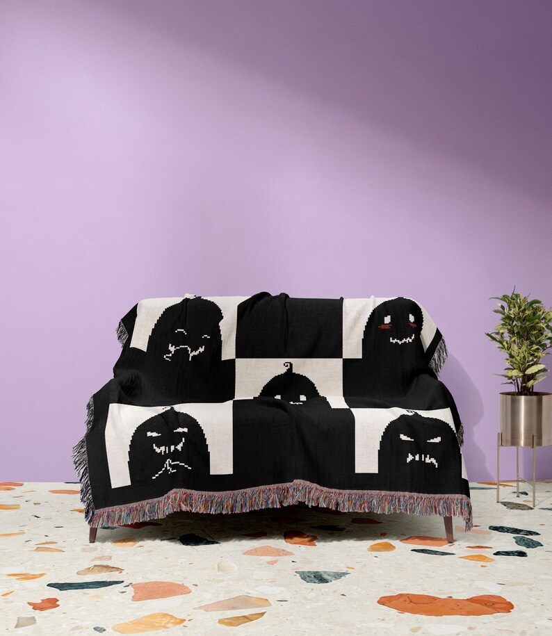 May include: Black and white woven throw blanket with a checkered pattern and a repeating design of black pumpkin faces. The blanket is draped over a black sofa with a fringe border.