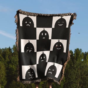 May include: A black and white woven throw blanket with a checkered pattern featuring various pumpkin faces. The blanket is held up by a person in a grassy field.