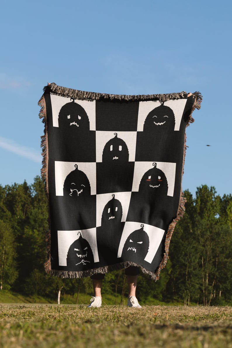 May include: A black and white woven throw blanket with a checkered pattern featuring various pumpkin faces. The blanket is held up by a person in a grassy field.