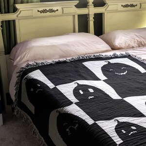 May include: A black and white woven throw blanket with a pattern of smiling ghosts. The blanket is draped over a bed with pink and white pillows.