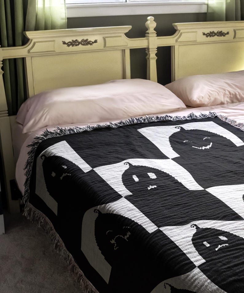 May include: A black and white woven throw blanket with a pattern of smiling ghosts. The blanket is draped over a bed with pink and white pillows.