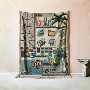 Peut inclure: Un plaid tiss&eacute; pr&eacute;sentant un design d'interface d'ordinateur des ann&eacute;es 90. Le motif inclut des ic&ocirc;nes pix&eacute;lis&eacute;es, un palmier, un poisson, un trombone anim&eacute;, ainsi que le texte "WordArt" et "Login successful" sur un fond beige.