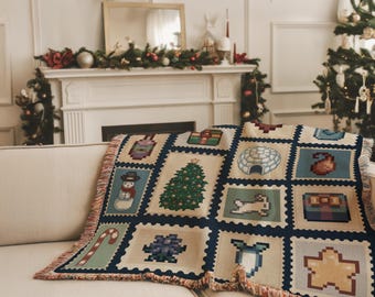 May include: A beige and blue Christmas-themed throw blanket with pixelated images of a snowman, Christmas tree, gifts, and other holiday icons. The blanket has a fringed edge and is draped over a cream-colored sofa.