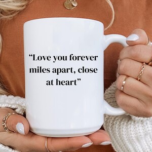 May include: A white ceramic mug with a curved handle. The mug features the text "Love you forever miles apart, close at heart" in black. The mug is held by a person wearing a white sweater.