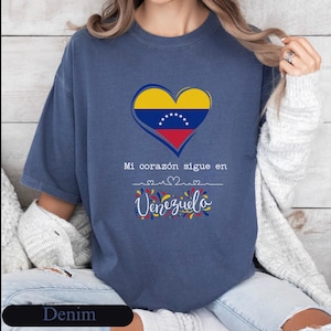 May include: A denim blue short-sleeve t-shirt featuring a heart graphic filled with the yellow, blue, and red colors of the Venezuelan flag. Below the heart, the text reads "Mi corazón sigue en" followed by "Venezuela" in script.