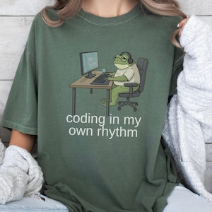 May include: Green t-shirt with the phrase "coding in my own rhythm." The shirt features a cartoon frog wearing headphones and glasses, sitting at a desk with a computer and keyboard. The frog is in a chair.