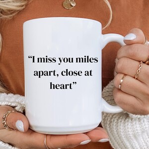 May include: White ceramic mug with a curved handle. The mug features the text "I miss you miles apart, close at heart" in black. The mug is held by a person wearing a white sweater and gold rings.