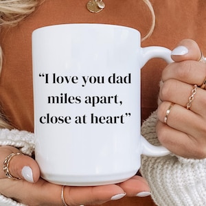 May include: A white ceramic mug with a curved handle. The mug features the text "I love you dad miles apart, close at heart" in black, centered on the front. The mug is held by a person wearing a white sweater.