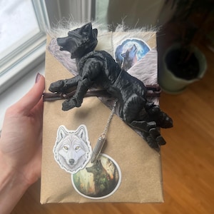 Könnte beinhalten: Ein verpacktes Geschenk mit einer schwarzen Wolfsfigur. Das Geschenk ist mit Wolfsaufklebern und einem silbernen Anhänger verziert. Die Verpackung ist braunes Papier mit Kunstfell und Bindfaden.