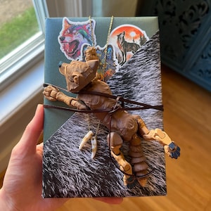 Puede incluir: Una caja de regalo rectangular envuelta en papel de color verde oscuro y estampado de piel negra, decorada con dos pegatinas de lobo, un cordón de cuero marrón, un collar dorado con dos colgantes de garra blancos y una figura de lobo articulada.