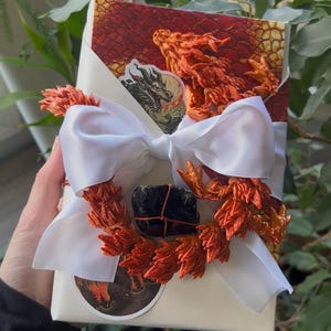Puede incluir: Un regalo envuelto con un lazo blanco y detalles decorativos naranjas. El papel de regalo presenta un patrón de escamas de dragón y una ilustración de dragón. Una piedra negra está atada al lazo.
