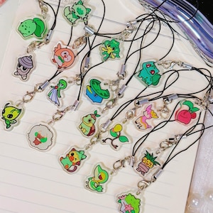 May include: A selection of colourful cartoon character charms, each attached to a black cord with a silver clasp. The charms are displayed on lined paper. The designs include various creatures.
