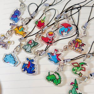 May include: A collection of colorful acrylic charms featuring various cartoon character designs. Each charm is attached to a black lanyard with a silver-tone clasp. The charms are displayed on a white notebook page.