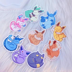 May include: A selection of vibrant cartoon stickers depicting various Pokémon characters. The stickers show different Pokémon in various poses, with a white border. The stickers are on a white, fluffy surface. The text "MINTY ARTZ" is visible on some stickers.