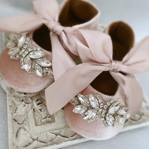 Peut inclure: Une paire de chaussures de b&eacute;b&eacute; en velours rose avec un grand ornement en strass sur chaque chaussure. Les chaussures sont attach&eacute;es ensemble par un n&oelig;ud en ruban de satin rose.