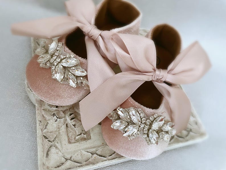 Peut inclure: Une paire de chaussures de b&eacute;b&eacute; en velours rose avec un grand ornement en strass sur chaque chaussure. Les chaussures sont attach&eacute;es ensemble par un n&oelig;ud en ruban de satin rose.