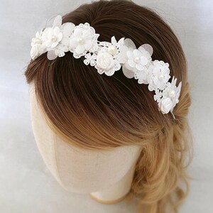May include: A white floral headband featuring delicate lace and pearl accents. The headpiece is adorned with fabric flowers and small pearls, creating an elegant and romantic design. Perfect for weddings or special occasions.