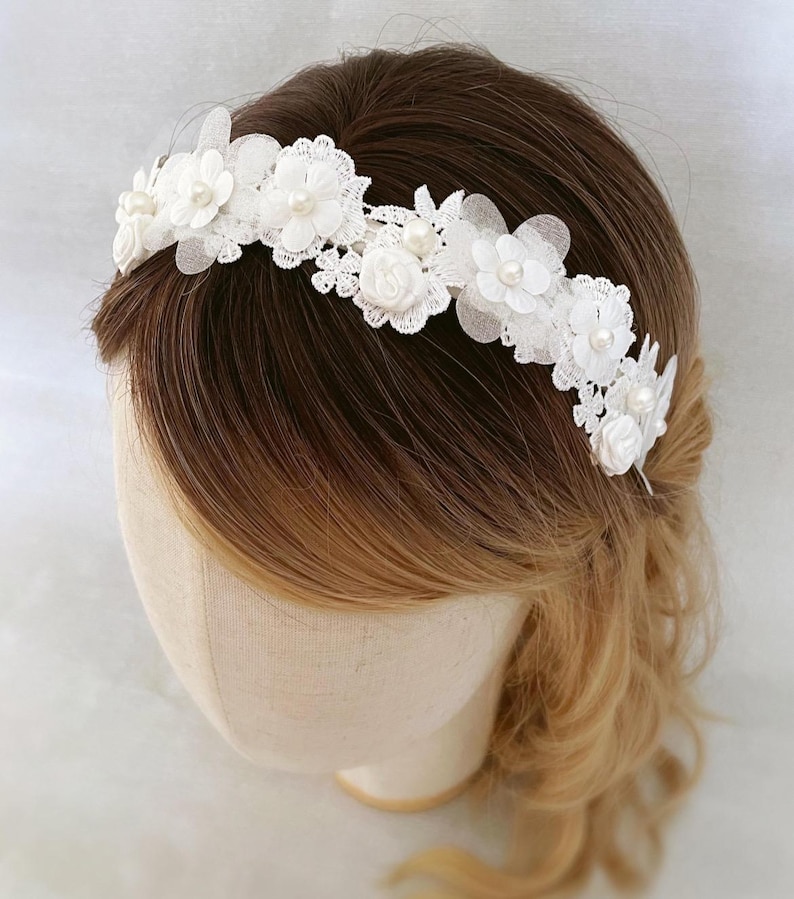 May include: A white floral headband featuring delicate lace and pearl accents. The headpiece is adorned with fabric flowers and small pearls, creating an elegant and romantic design. Perfect for weddings or special occasions.
