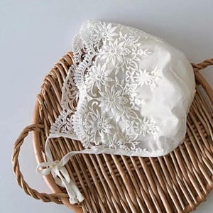 May include: A white lace baby bonnet with a floral design. The bonnet is tied with a white ribbon and is sitting on a wicker basket.
