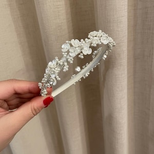 May include: A white headband adorned with delicate white flowers and small, round pearls. The floral embellishments are clustered along the top of the band, creating a decorative accent. The headband is held against a neutral-toned background.