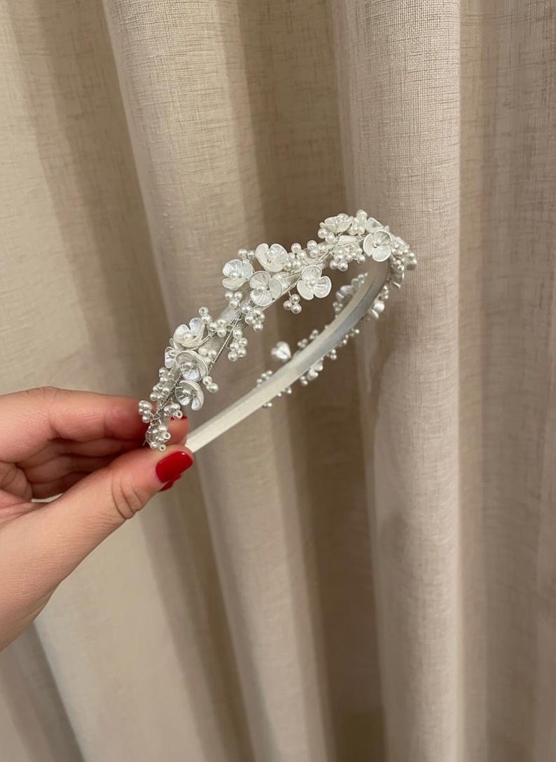 May include: A white headband adorned with delicate white flowers and small, round pearls. The floral embellishments are clustered along the top of the band, creating a decorative accent. The headband is held against a neutral-toned background.