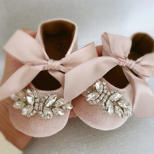 Peut inclure: Une paire de chaussures de b&eacute;b&eacute; en velours rose avec un n&oelig;ud en satin et un grand embellissement en strass.