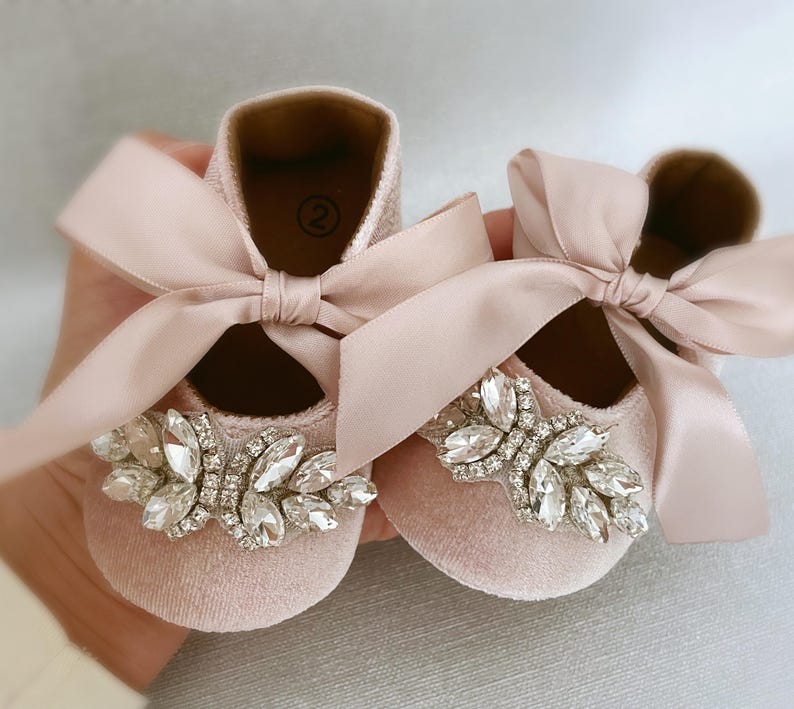 Peut inclure: Une paire de chaussures de b&eacute;b&eacute; en velours rose avec un n&oelig;ud en satin et un grand embellissement en strass.