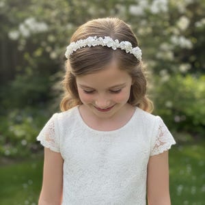 May include: A young person wearing a white lace dress and a floral crown. The crown is made of small white flowers and beads. The dress has short sleeves and a round neckline. The background is a blurred view of green foliage.