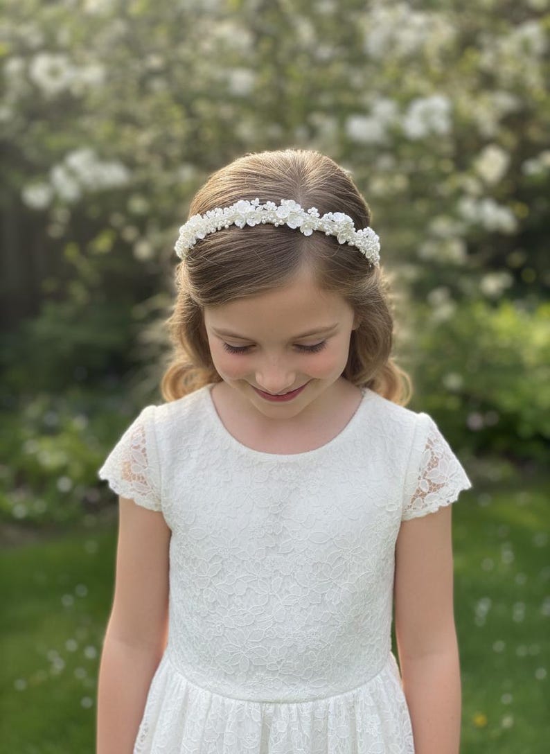 May include: A young person wearing a white lace dress and a floral crown. The crown is made of small white flowers and beads. The dress has short sleeves and a round neckline. The background is a blurred view of green foliage.