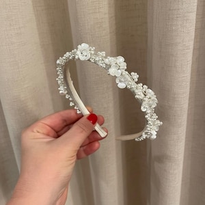 May include: A white headband adorned with small white flowers and faux pearls. The floral and pearl embellishments are clustered along the top of the headband, creating a delicate and elegant design. Perfect for weddings or special occasions.