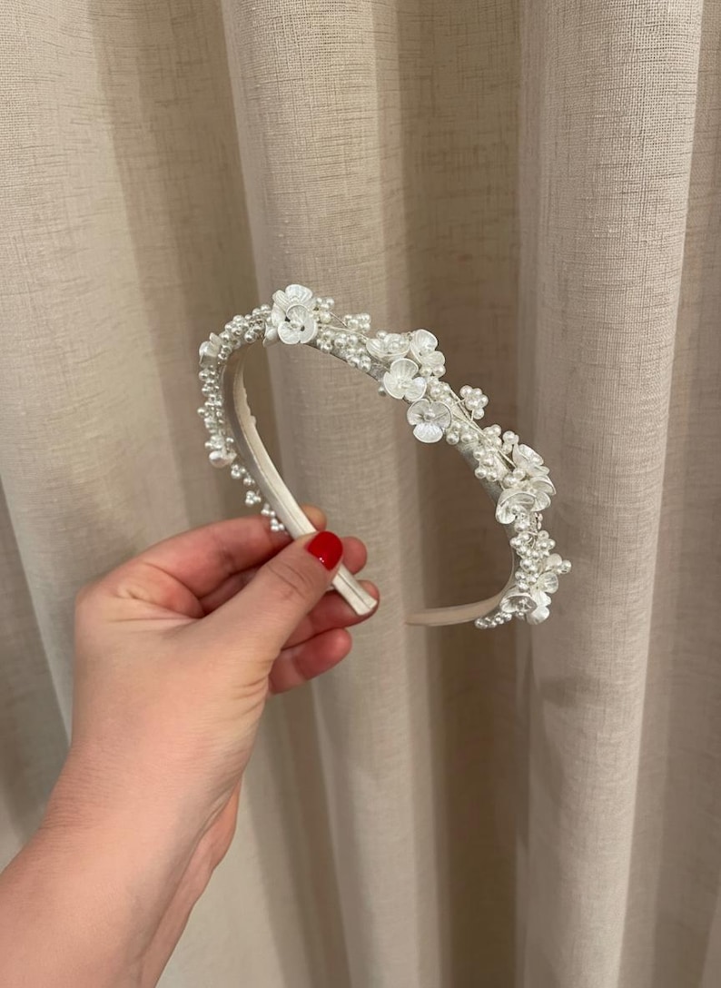 May include: A white headband adorned with small white flowers and faux pearls. The floral and pearl embellishments are clustered along the top of the headband, creating a delicate and elegant design. Perfect for weddings or special occasions.