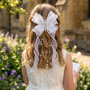 Op de afbeelding: Een witte organza haarstrik met een kruisvormige versiering en bungelende imitatieparels. De strik is bevestigd in het haar van een jong persoon, passend bij een witte jurk. Geschikt voor een speciale gelegenheid.
