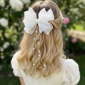 Peut inclure: Un nœud de cheveux blanc en tissu transparent, orné d'un centre de perles et de brins de perles en cascade. Le nœud est placé à l'arrière de la tête, avec des cheveux blonds visibles. L'arrière-plan montre un jardin flou avec des fleurs blanches et roses.