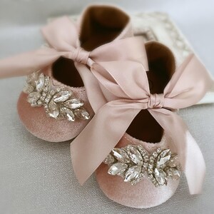 Peut inclure: Une paire de chaussures pour b&eacute;b&eacute; en velours rose avec un grand ornement en strass et un n&oelig;ud en satin rose assorti.
