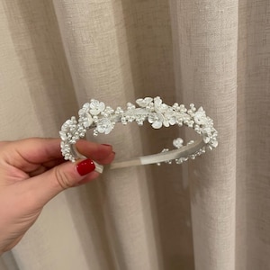 May include: A delicate white floral headband with pearl accents. The headband features small, fabric flowers and clusters of pearls, creating a romantic and elegant design. Perfect for weddings or special occasions.