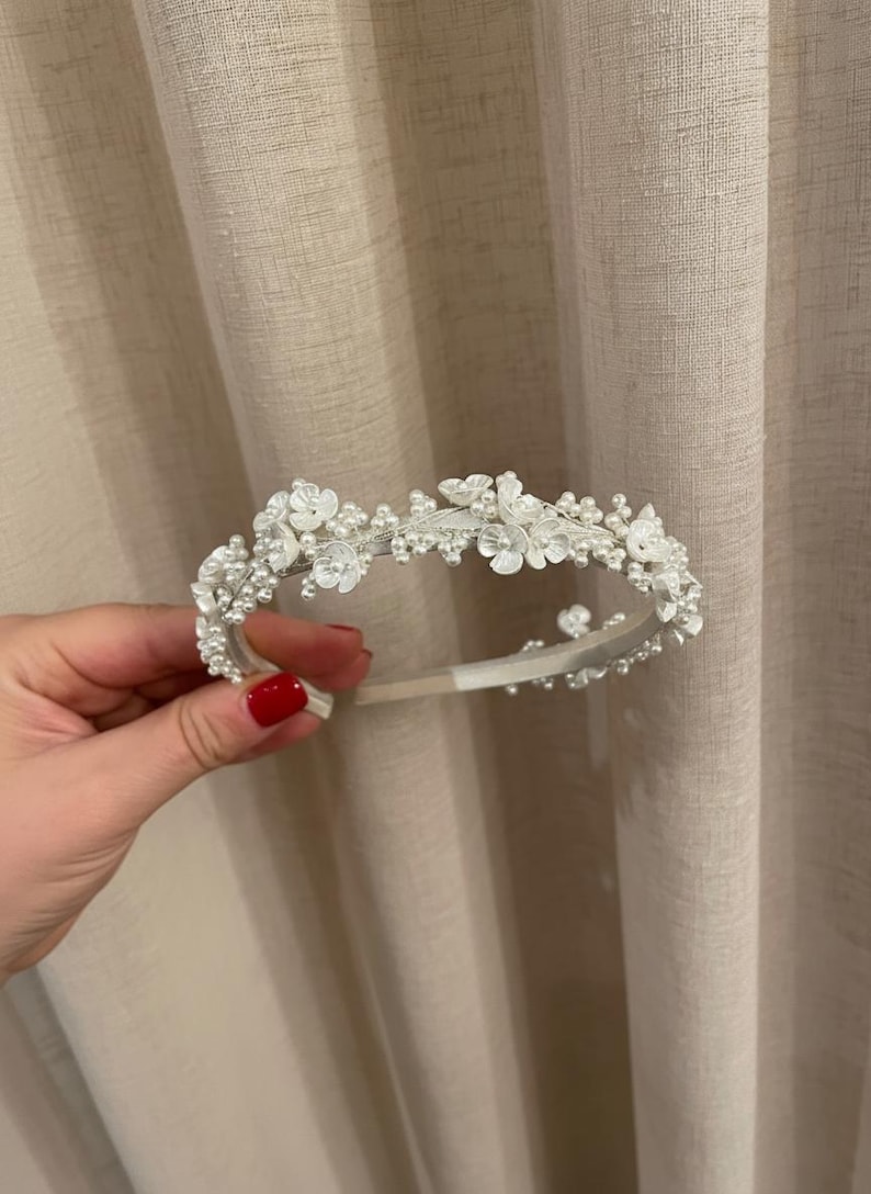 May include: A delicate white floral headband with pearl accents. The headband features small, fabric flowers and clusters of pearls, creating a romantic and elegant design. Perfect for weddings or special occasions.