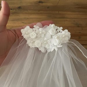 May include: A white tulle veil with a floral headpiece. The headpiece features small white fabric flowers and faux pearls. The veil is made of a sheer, lightweight fabric, suitable for weddings or special occasions.
