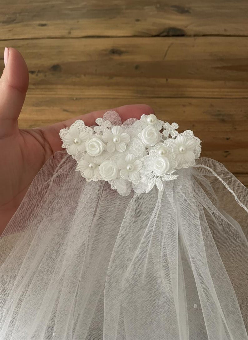 May include: A white tulle veil with a floral headpiece. The headpiece features small white fabric flowers and faux pearls. The veil is made of a sheer, lightweight fabric, suitable for weddings or special occasions.