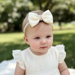 May include: A baby wearing a cream-coloured bow headband with a rhinestone centre. The headband is paired with a cream-coloured dress with eyelet detailing and ruffled sleeves. The background is a blurred green outdoor scene.