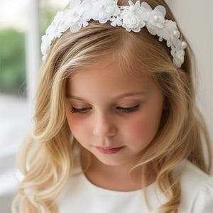 May include: A young girl with long, wavy blonde hair wearing a white floral headband with pearl accents. The headband features delicate fabric flowers and small pearls. She is wearing a white dress.