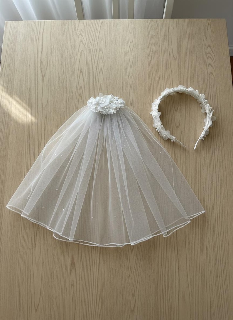 May include: A white bridal veil with a floral embellishment and a matching floral headband. The veil is made of sheer fabric and has a rounded shape. The headband is a simple band adorned with small white flowers.
