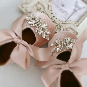 Peut inclure: Une paire de chaussures de b&eacute;b&eacute; en velours rose poudr&eacute; avec un grand embellissement en strass sur chaque chaussure. Les chaussures sont orn&eacute;es d'un n&oelig;ud en satin.