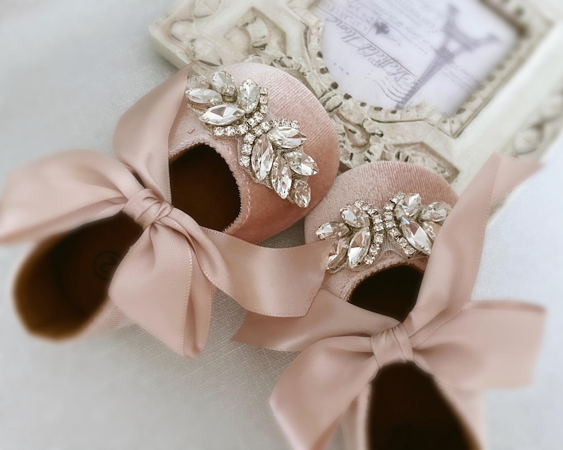 Peut inclure: Une paire de chaussures de b&eacute;b&eacute; en velours rose poudr&eacute; avec un grand embellissement en strass sur chaque chaussure. Les chaussures sont orn&eacute;es d'un n&oelig;ud en satin.