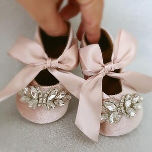 Peut inclure: Une paire de chaussures pour b&eacute;b&eacute; avec des n&oelig;uds en satin rose et des embellissements en cristal. Les chaussures sont faites d'un tissu rose doux et ont un design d&eacute;licat et f&eacute;minin.